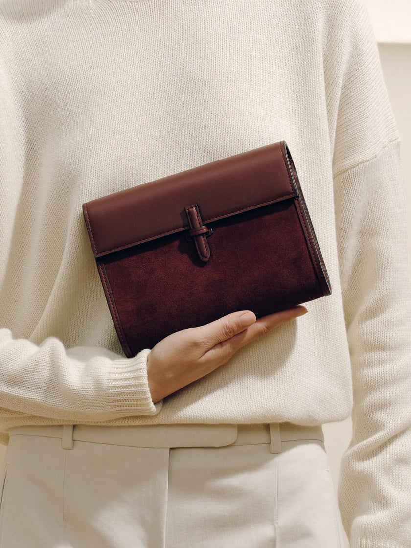 Brown leather clutch held by a person wearing a beige sweater and pants.